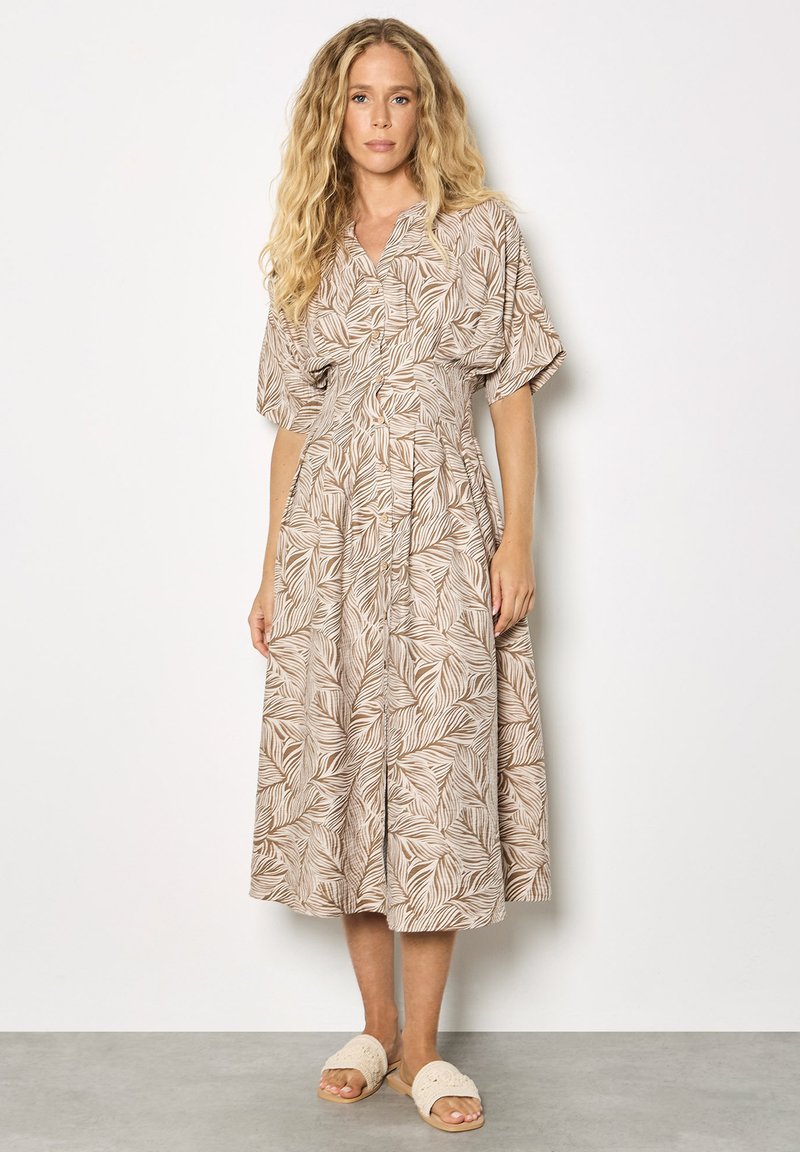 Femme aux longs cheveux blonds portant une robe midi beige et blanche à motif de feuilles et des sandales beiges à enfiler, debout contre un mur blanc.