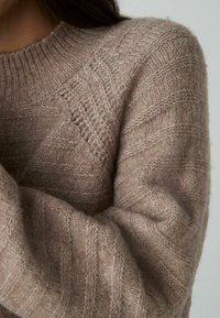 Jasnobrązowy sweter dzianinowy o teksturowanym wzorze, z ribbowanym kołnierzem i rękawami, wykonany z miękkiego, puszystego materiału.