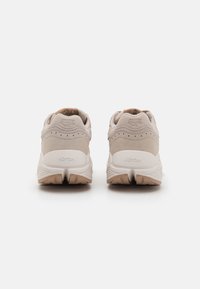Beige színű sportcipők, velúr és hálós felsőrész, párnázott cipőgallér, Vibram talp és texturált részletek; a kényelem és a támogatás érdekében tervezve.