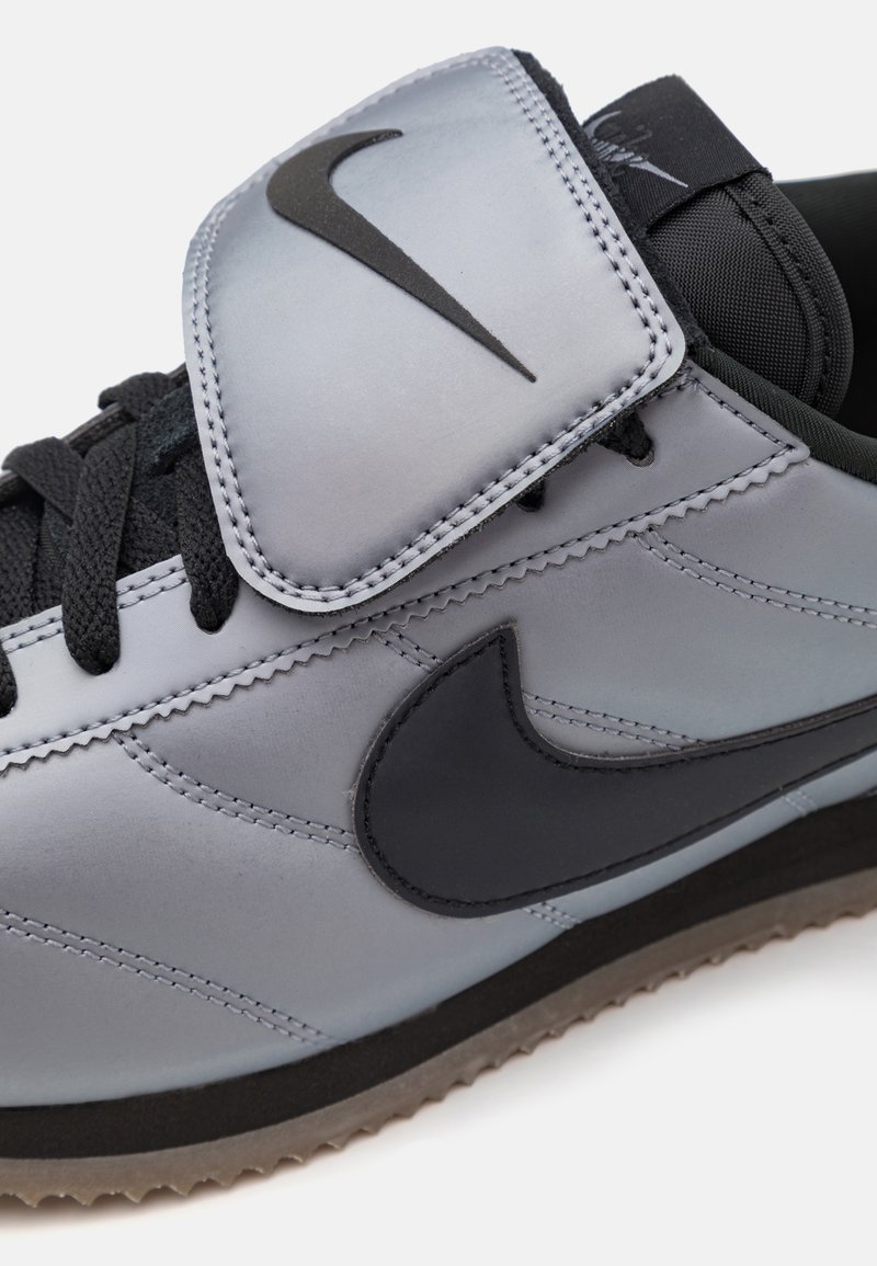 Zapatilla deportiva gris con un upper sintético suave, que presenta un gran swoosh negro, cordones negros y una correa de velcro para el cierre.