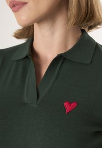 Donkergroen poloshirt met een kraag, voorzien van een kleine rode hartborduursel aan de linkerkant. Glad materiaal, casual ontwerp.