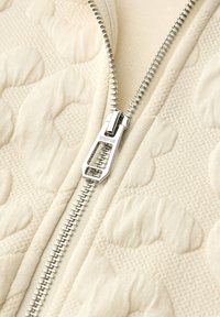 Primo piano di una cerniera in metallo argento su tessuto trapuntato crema con motivi in rilievo e cuciture testurizzate.