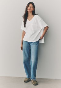 White short-sleeve V-neck shirt with a pocket, paired with loose-fit blue denim jeans and brown clogs, set against a light background.