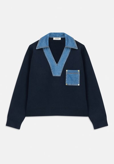 Felpa navy con scollo a V profondo, dotata di colletto e tasca in denim. Il tessuto appare morbido, con una vestibilità rilassata e cropped.