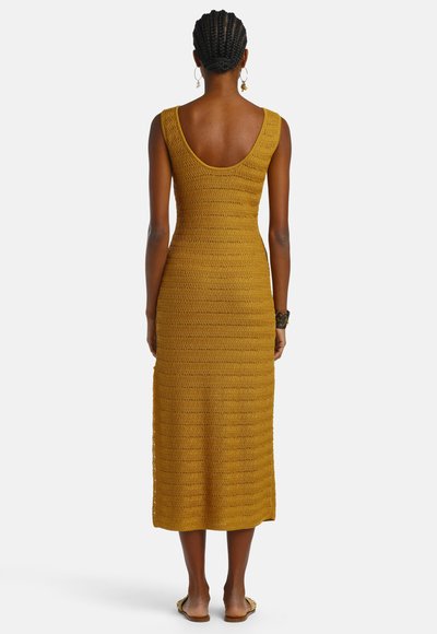 Femme aux cheveux tressés portant une robe midi sans manches en maille jaune moutarde, boucles d'oreilles créoles, bracelet et sandales plates, debout de dos.