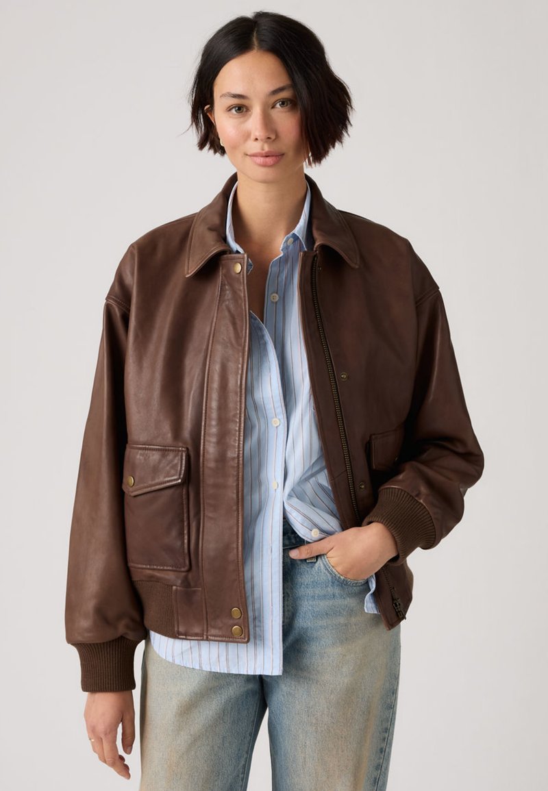 Jauna moteris su trumpais tamsiais plaukais, vilkinti rudą odinę striukę, mėlyną dryžuotą marškinius ir šviesiai mėlynas džinsines kelnes, stovinti neutralaus fono apsuptyje.