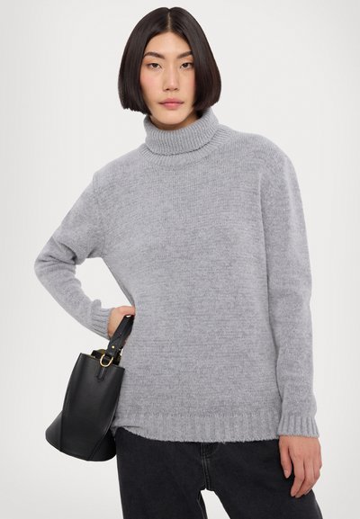 Pulover gri din tricot cu guler înalt ribanat, croială lejeră și manșete ribanate. Asortat cu o geantă din piele neagră, pe un fundal simplu.