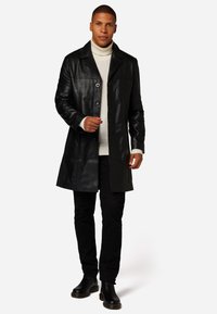 Schwarzer Ledermantel mit Knopfverschluss, maßgeschneiderter Schnitt und langen Ärmeln; getragen über einem hellen Pullover und kombiniert mit dunklen Hosen und Stiefeln.