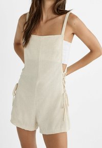 Leinen-Jumpsuit in hellbeige mit verstellbaren Bändern an den Seiten, quadratischem Ausschnitt, breiten Trägern und einer lockeren Passform. Glatte Textur überall.