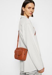 Borsa a tracolla in pelle marrone con pattina frontale, hardware dorato e tracolla regolabile. La borsa presenta una superficie testurizzata e una forma rettangolare.