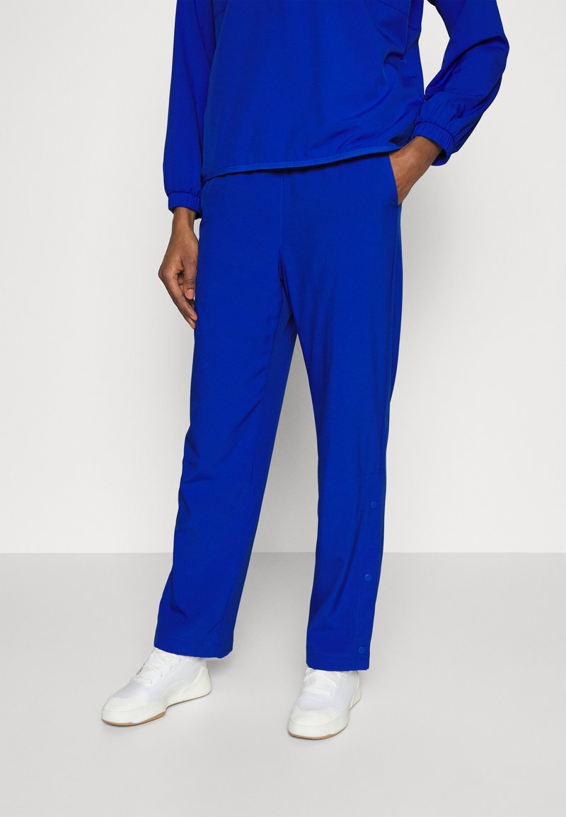 Blauwe sportbroek van lichtgewicht stof, met een losse pasvorm en drukknoopsluiting aan de zijkant. Draagt wit sneakers.