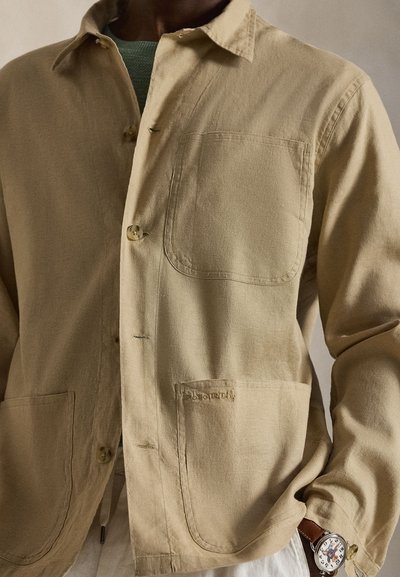 Άνδρας που φοράει μπεζ σακάκι με κουμπιά και μεγάλες μπροστινές τσέπες, από πάνω ενός πράσινου πουκαμίσου, και ένα ρολόι χειρός με καφέ δερμάτινο λουρί.