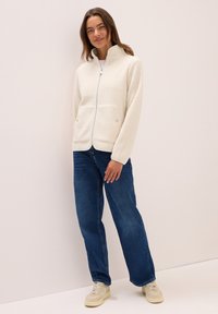 Femme portant une veste en polaire crème zippée, un jean bleu à jambes larges et des baskets beiges, se tenant devant un fond clair.