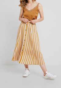 Camiseta sin mangas color tostado con borde de encaje combinada con una falda midi de rayas verticales en crema y mostaza con detalles de botones negros. Zapatillas blancas.