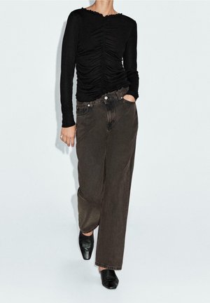 Person wearing a ruched black long-sleeve top, loose dark gray jeans, and black square-toe shoes standing against a light background.