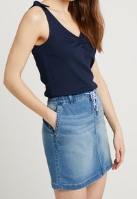 Marineblaues geripptes Tanktop mit V-Ausschnitt und Schulterbändern; kombiniert mit einem himmelblauen Jeansrock mit Bindegürtel und Taschen.