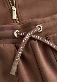 Bruine joggers met een gladde textuur, voorzien van gouden hardware en trekkoordjes met de logo's "BAKER" en "TED BAKER" in het wit.