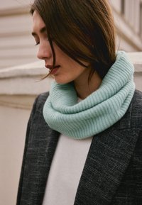 A soft, ribbed cowl neck scarf in light mint green, worn with a gray blazer over a white top, highlighting the cozy texture and layering.