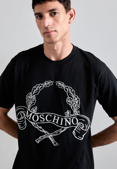Μαύρο βαμβακερό μπλουζάκι με λευκό γραφικό σχέδιο από δάφνινα φύλλα και τη λέξη "MOSCHINO" σε ένα κορδόνι στη μέση μπροστά.