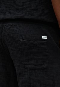 Κοντινή λήψη τσέπης σε μαύρο υφασμάτινο παντελόνι με υφή, με λευκή ετικέτα της μάρκας Jack & Jones στη δεξιά πλευρά του γοφού.