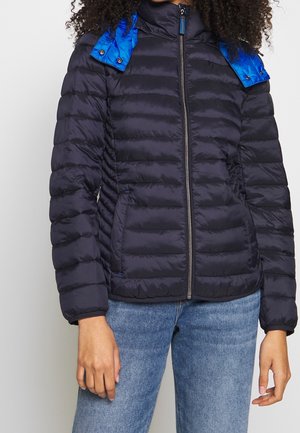 Frau trägt eine schwarze Steppjacke mit glänzenden blauen Akzenten am Kragen und blaue Jeans, steht mit entspannt an den Seiten hängenden Armen.