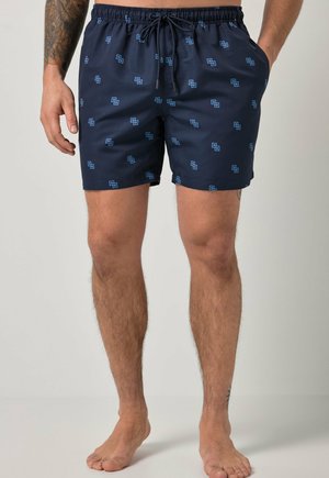 Mand iført marineblå badeshorts med lyseblåt geometrisk mønster, snøre i taljen, hænderne i lommerne, stående barfodet.