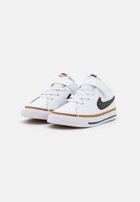 White sneakers with a black swoosh, orange and black accents, rubber sole, and textured detail on the upper. Velcro strap and laces included.