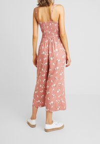 Frau in einem ärmellosen, rosa Blumen-Overall mit gerafftem Oberteil und weißen lässigen Turnschuhen, die vor einem schlichten, hellen Hintergrund steht.