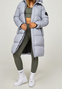 Hellgrauer gepolsterter langer Mantel mit Kapuze, durchgehendem Reißverschluss, schwarzem Logo-Patch, kombiniert mit olivgrünem, figurbetontem Oberteil und Leggings sowie weißen Turnschuhen.