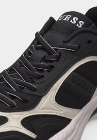 Ανδρικό αθλητικό παπούτσι ασπρόμαυρο με κορδόνια με σχέδιο, πλέγμα στη γλώσσα που δείχνει μερικό κείμενο λογότυπου, και διαφοροποιημένο σχέδιο στην σόλα.