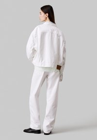 Chaqueta de mezclilla blanca de corte oversize, con costuras visibles y una etiqueta rectangular marrón. Combinada con pantalones blancos de corte holgado a juego.