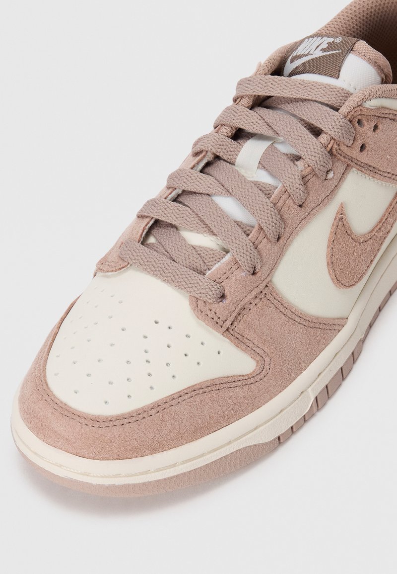 Sneaker Nike low-top con sovrapposizioni in suede beige, pannelli in pelle crema, punta perforata e stringhe beige abbinate su sfondo bianco.