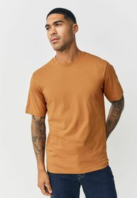 Īsām piedurknēm T-krekls siltā brūnā krāsā, izgatavots no gluda kokvilnas materiāla, ar apaļu kaklu un brīvu piegriezumu. Parādīts kopā ar ziliem džinsiem.