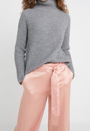 Femme portant un pull à col roulé gris côtelé et un pantalon en satin rose clair avec une ceinture ruban nouée, les mains dans les poches.