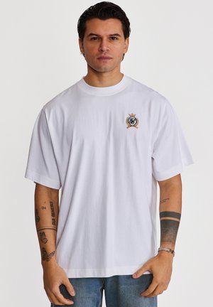 Homme portant un t-shirt blanc oversize avec un petit blason brodé sur la poitrine, un jean bleu, et des tatouages visibles sur les deux avant-bras.