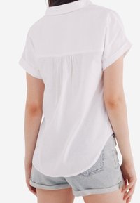 Chemise blanche à manches courtes avec col, présentant un détail plissé au dos et un ourlet arrondi, associée à un short en denim clair.