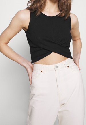Woman wearing a black sleeveless crop top with a twist front, paired with high-waisted light beige pants, hands resting on hips.