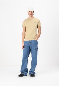 Beige, testhezálló póló, kék cargo farmer, oldalzsebbel, és fekete slip-on cipő. A modellen fehér sapka van, és egy fehér háttér előtt áll.