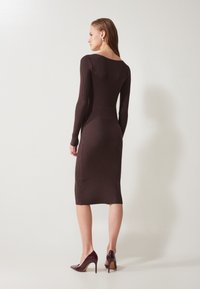 Brown ribbed knit form-fitting dress with long sleeves, side slit detailing, and a scoop neckline, paired with glossy burgundy high heels.