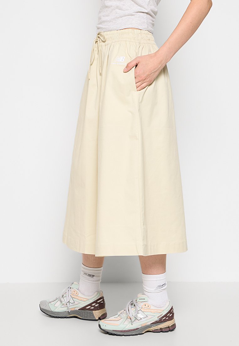 Falda midi beige claro hecha de algodón con una cintura elástica, bolsillos laterales y un detalle de lazo. Complementada con zapatillas grises y pasteles.