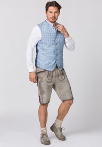 Hellblaues gemustertes Midivest über einem weißen Hemd, kombiniert mit grauen Ledershorts, die dekorative Stickereien und Knopfdetails aufweisen.
