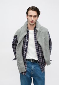 Szürke kötött kardigán sötétkék és piros kockás ing fölött, fehér pólóval és kék farmernadrággal párosítva. Fekete övvel együtt.