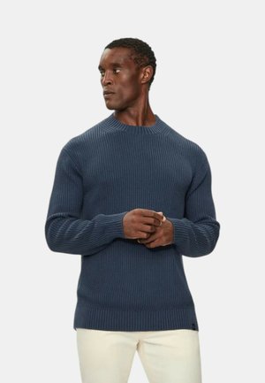 Maglione a coste blu navy con scollatura tonda, maniche lunghe e tessuto strutturato; abbinato a pantaloni di colore chiaro.