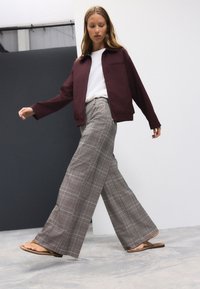 Giacca bordeaux, T-shirt bianca, pantaloni ampi a quadri grigi e infradito marroni, in piedi su un pavimento di cemento contro uno sfondo neutro.