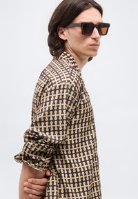 Άνδρας με μεσαίου μήκους σκούρα μαλλιά, φοράει μεγάλα τετράγωνα γυαλιά ηλίου και καφέ πουκάμισο με μοτίβο και μακριά μανίκια, στέκεται με τα χέρια πίσω από την πλάτη.