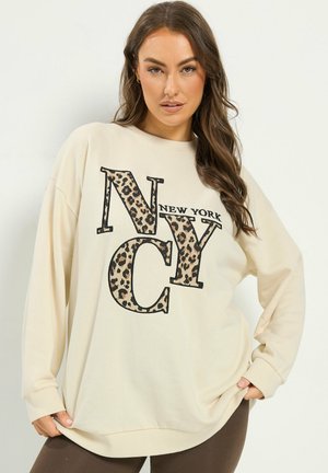 Femme portant un sweat crème avec de grandes lettres imprimées léopard "NYC" et "New York", posant avec les mains tenant les côtés du sweat.