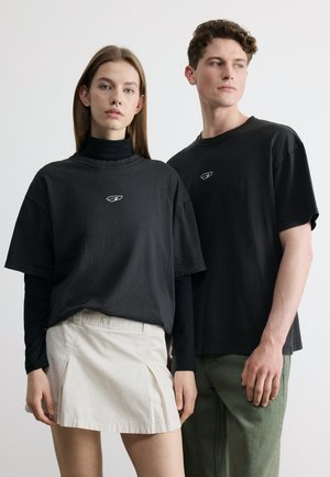 Jeune femme et homme portant des hauts noirs avec de petits logos blancs, debout devant un fond clair uni, regardant vers l'avant et de profil.