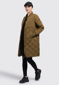 Veste longue matelassée vert olive avec poches latérales, poignets côtelés et fermeture éclair à la base ; portée sur un haut noir et un pantalon ajusté avec des baskets noires.