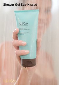 AHAAVA Sea-Kissed Duschgel i en turkos tub med en brun bas, som visar produktinformation, hålls under rinnande vatten.