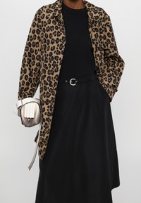 Leopardmönstrad jacka i beige och brun, bärs över en svart topp och svart kjol, håller en krämfärgad och silverfärgad handväska med veckad detalj.